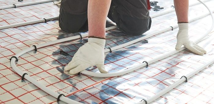 Person kniet auf Boden und verlegt Heizungsrohre auf einer Unterlage mit rotem Rastermuster, trägt weiße Handschuhe.