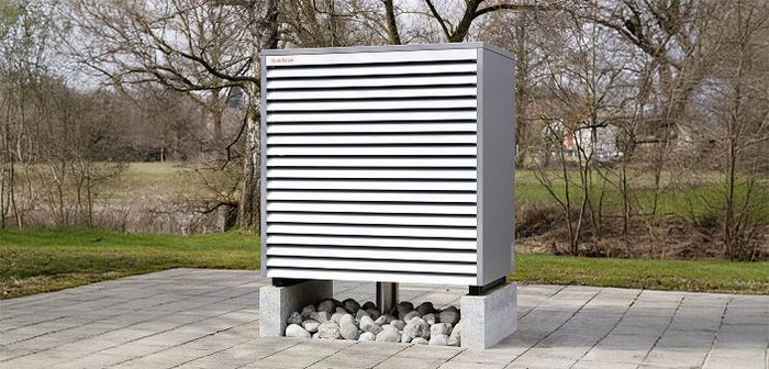 Wetterstation mit lamellenverkleidetem Messgerät auf Steinsockel im Freien, umgeben von Bäumen und Wiese.